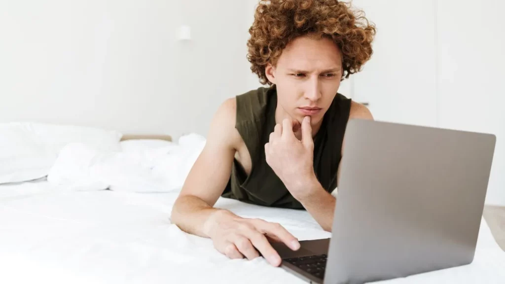 Man with curly hair lying on a bed using a laptop.