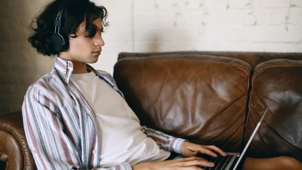 Man with headphones sitting on a leather sofa using a laptop.