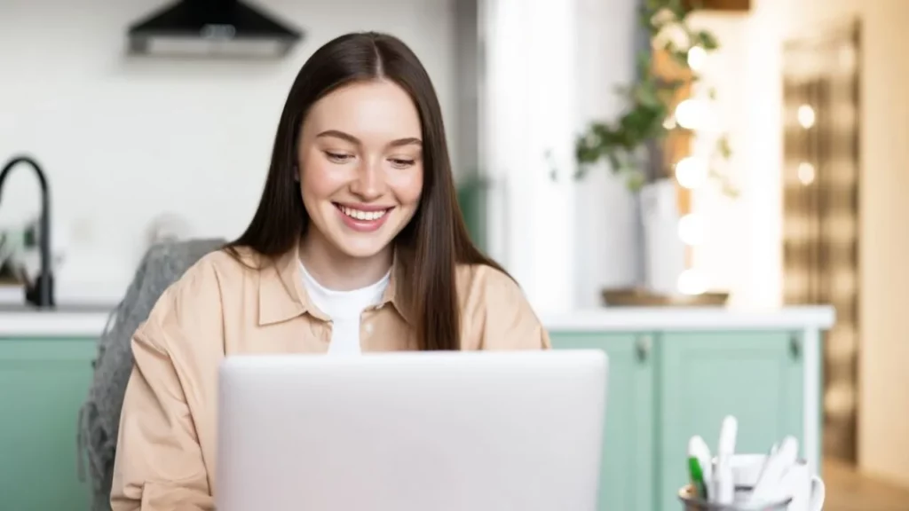 Happy young woman with long brown hair working on her laptop in a bright kitchen.