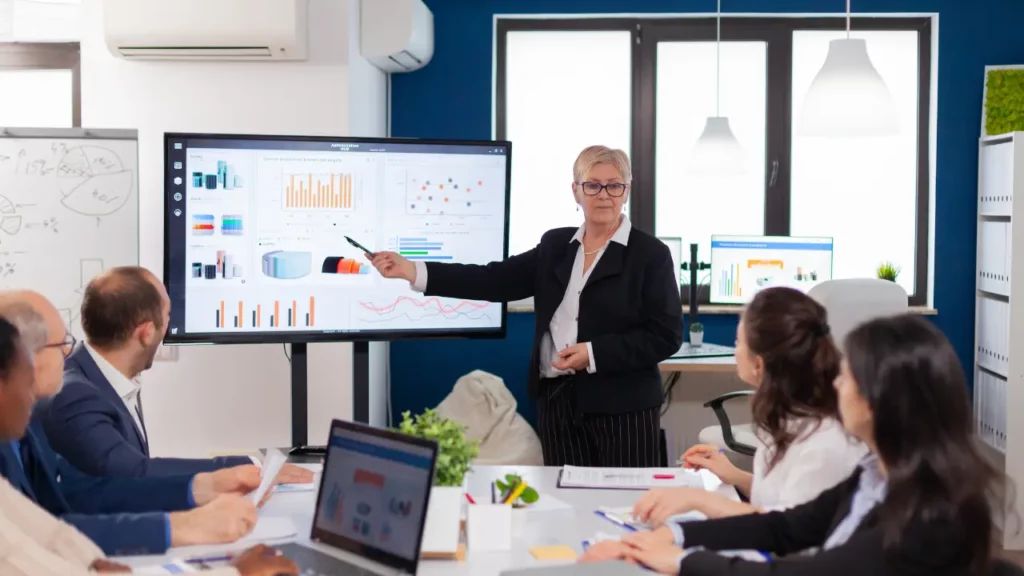 A woman in a business suit presenting data charts on a large monitor to a group of colleagues in a conference room.