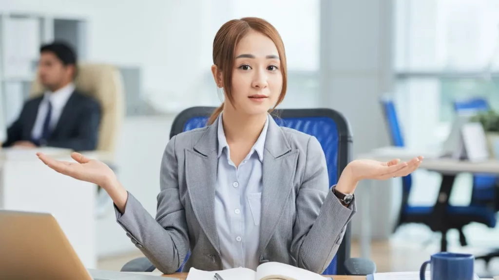 A woman in a grey business suit sitting at a desk and shrugging with both hands open in a confused or uncertain gesture.