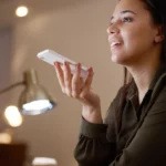 A woman smiling while holding a white smartphone horizontally near her mouth to record a voice message or use a voice assistant in front of a computer monitor.
