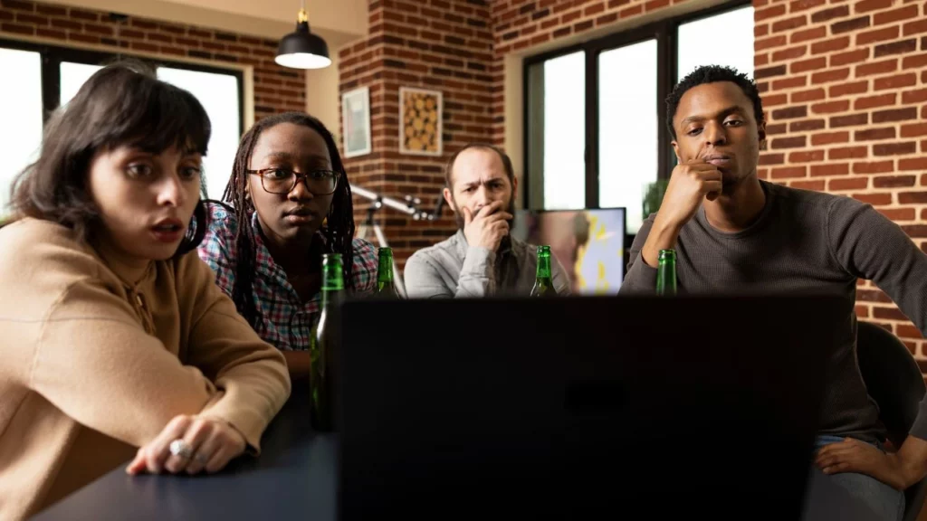 A diverse group of four individuals gathered together looking intently and with shocked or serious expressions at a laptop screen in a room with a brick wall.