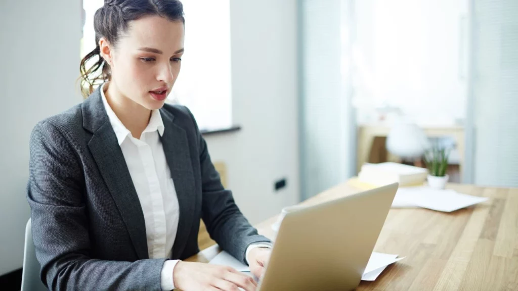 A woman in a grey business suit sitting at a wooden desk and typing on a laptop computer.
