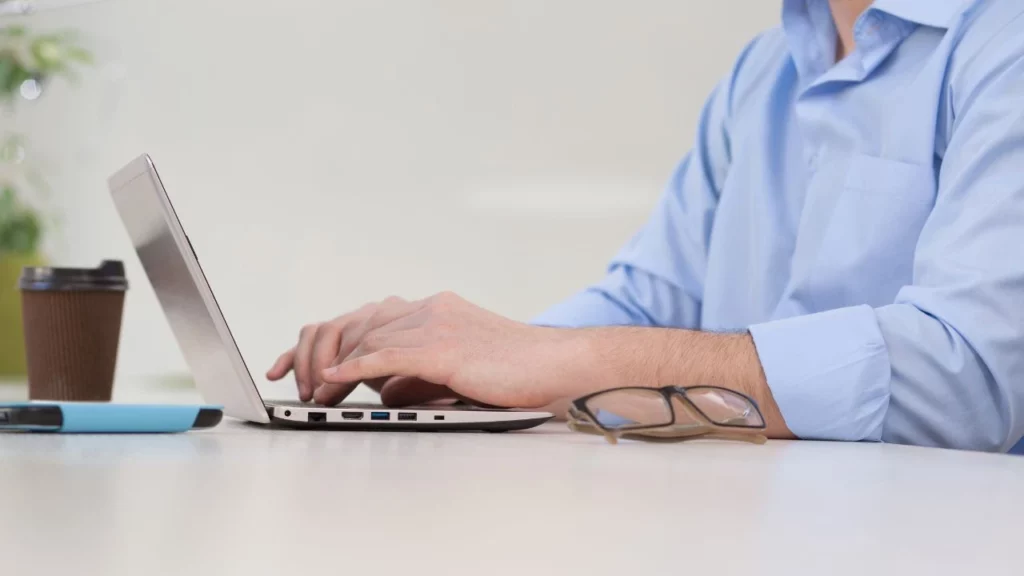 A close-up side view of a man in a light blue button-down shirt typing on a laptop at a desk with a pair of eyeglasses next to it.