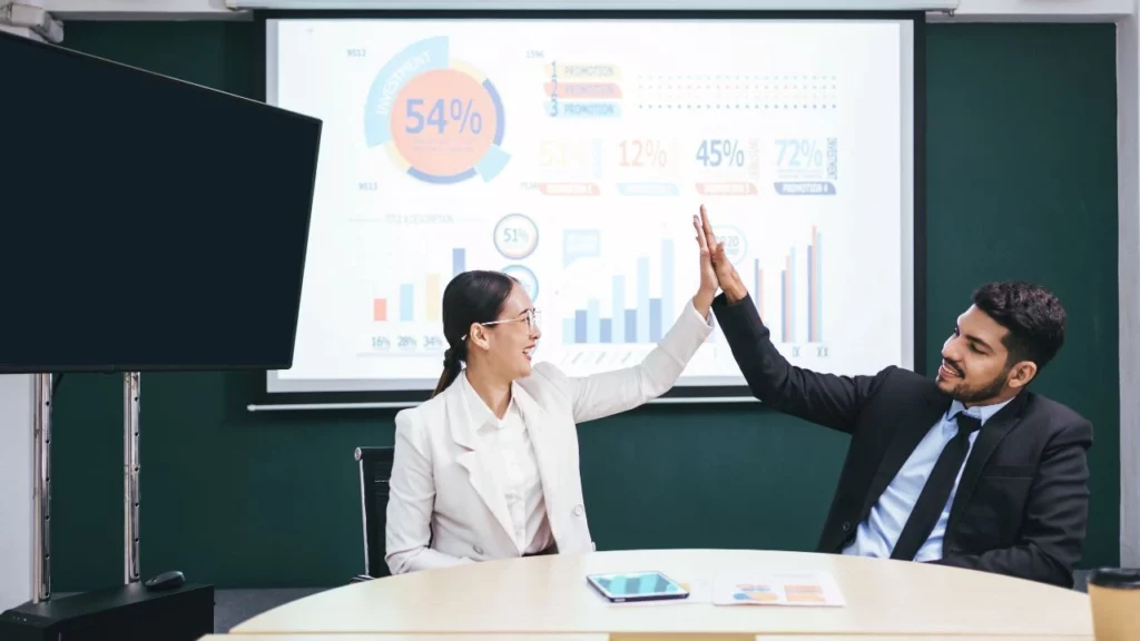 A man and a woman in business attire giving each other a high-five at a conference table, with a projector screen showing data charts and graphs behind them.